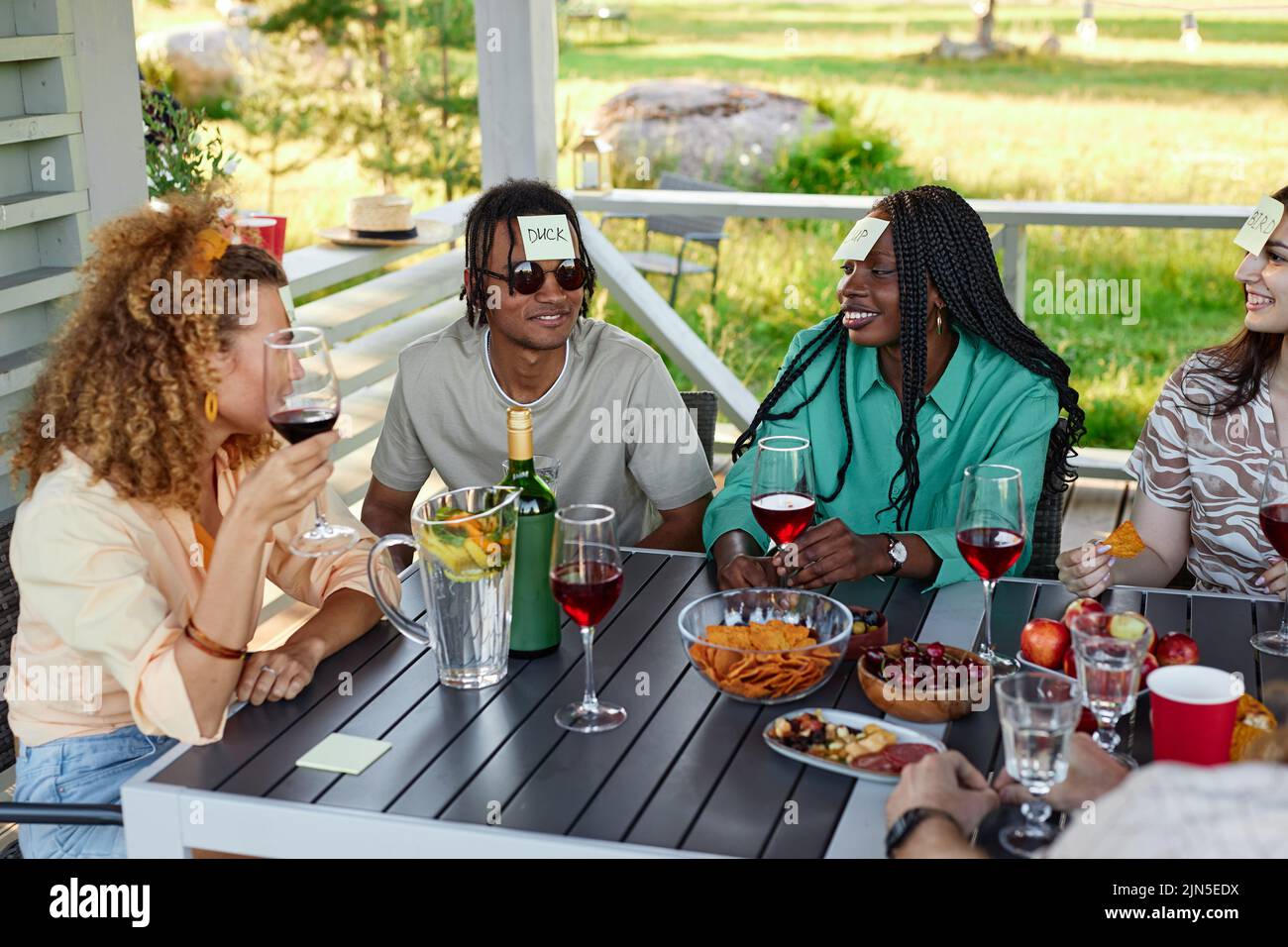 Diverse group of young people playing Guess who game while sitting at table outdoors in Summer Stock Photo