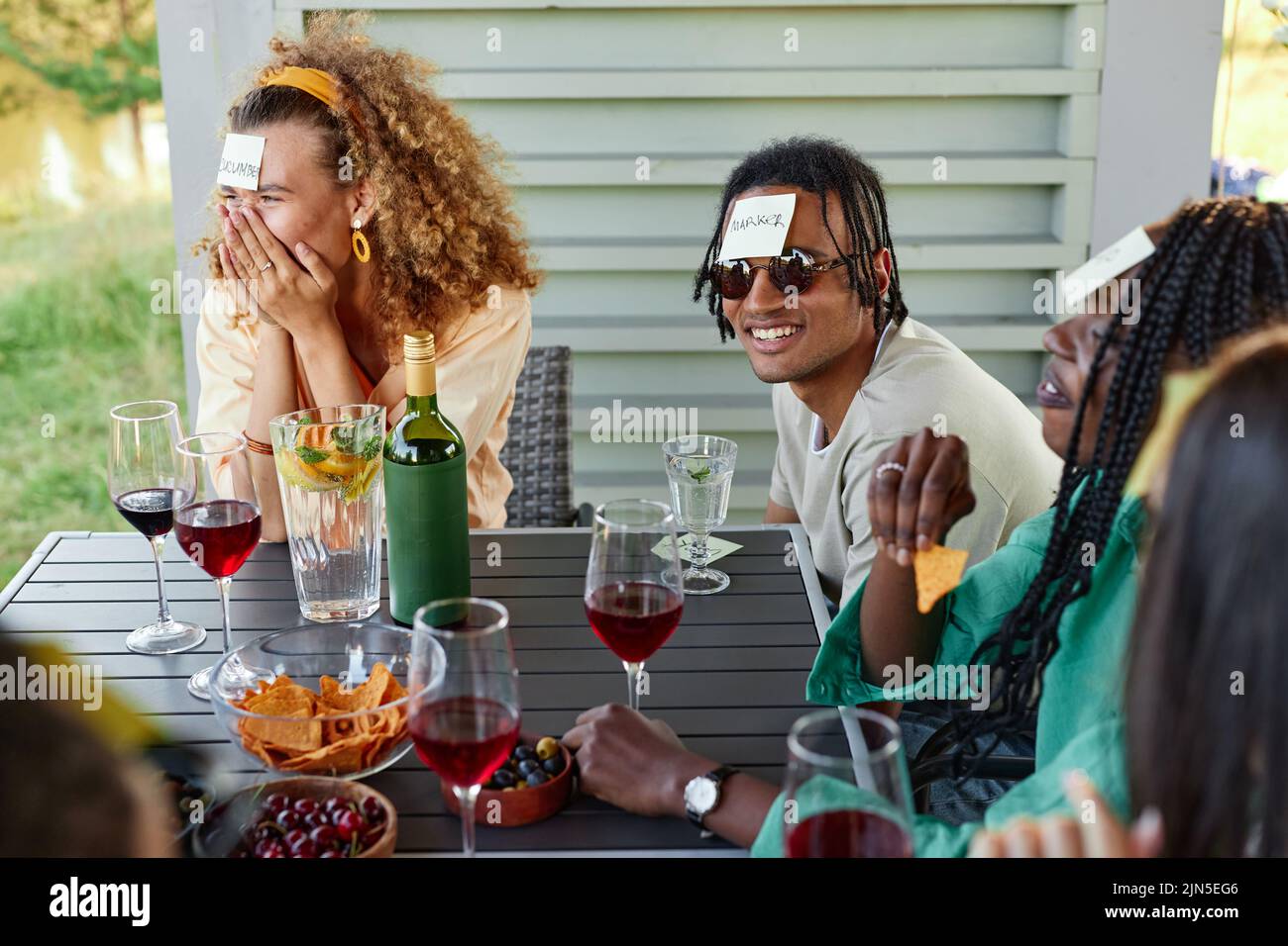 Group of young people laughing and playing Guess who game while sitting at table outdoors in Summer Stock Photo