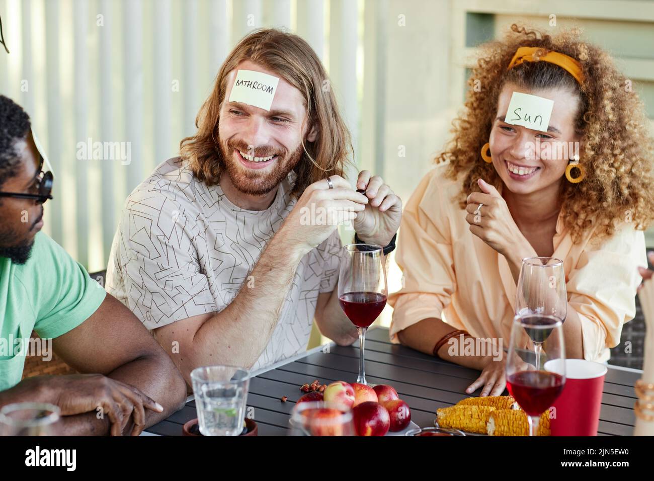 Portrait of smiling man with sticker note on head playing Guess who game with group of friends at outdoor party Stock Photo