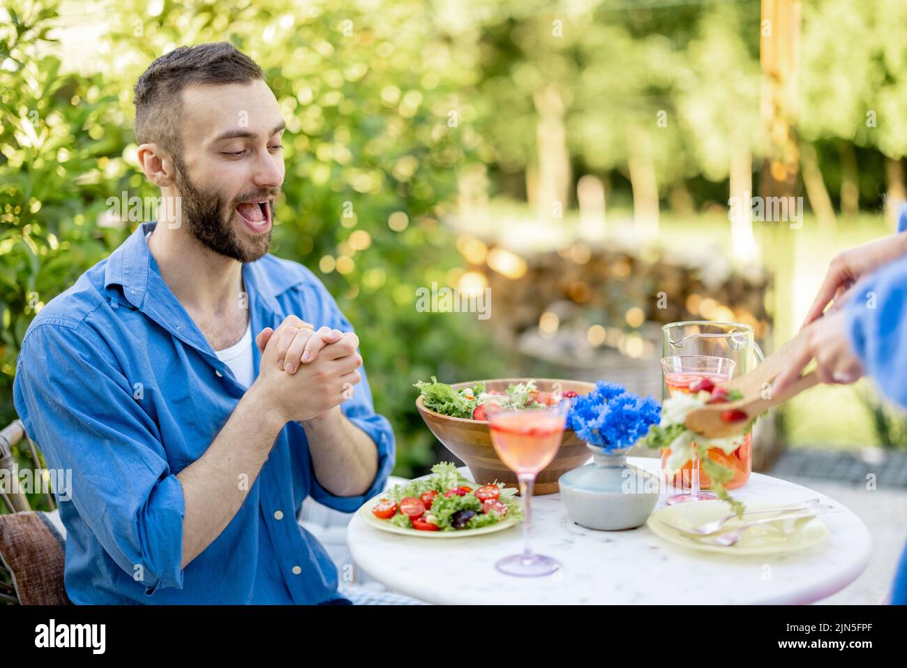Excited man is waiting for lunch, watching enthusiastically as his wife serves healthy salad. Young family have a lunch at backyard during summer time Stock Photo
