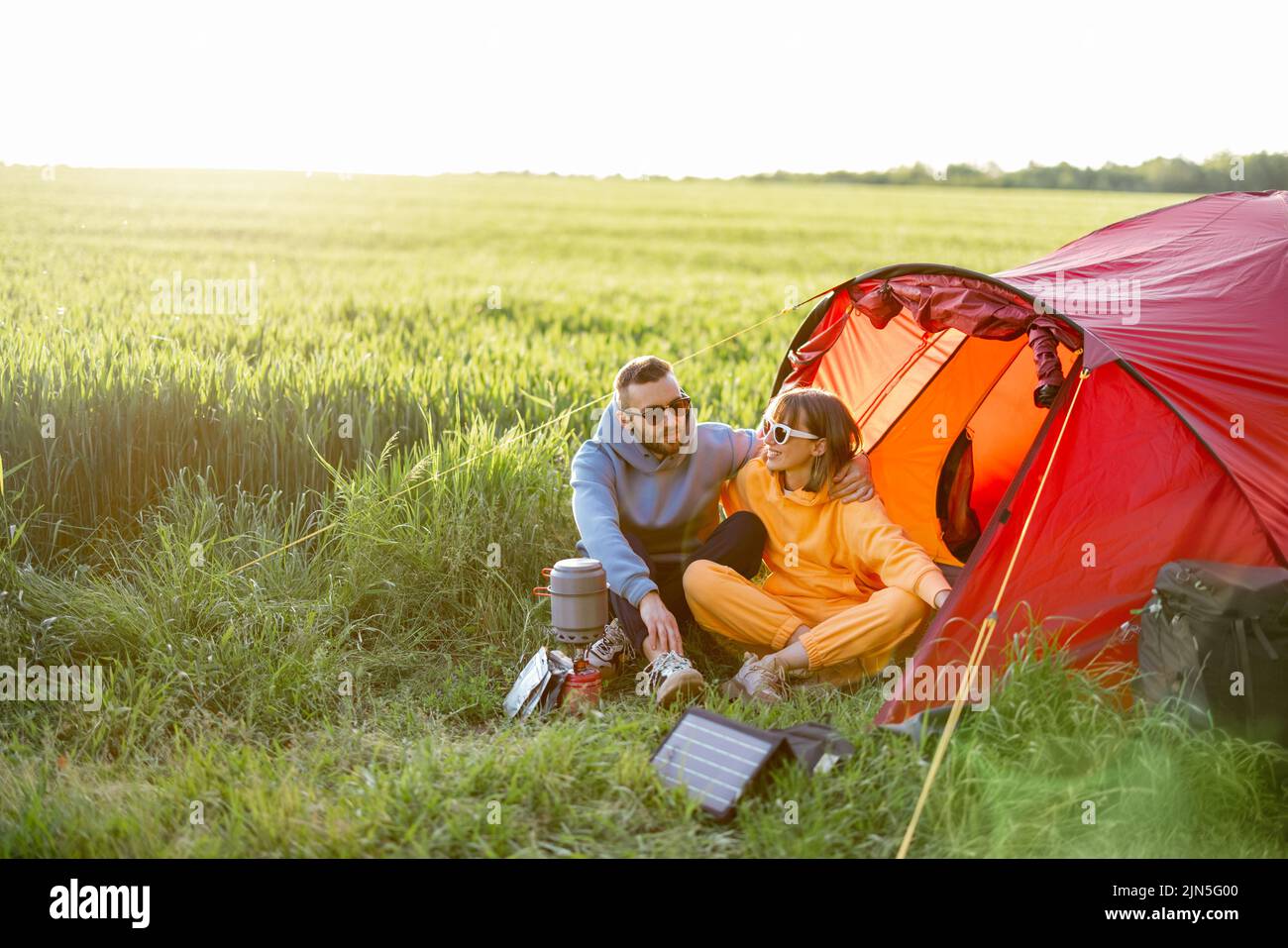 Young couple spending summer time together at campsite near tent on the green field. Charging gadgets with portable solar panel Stock Photo