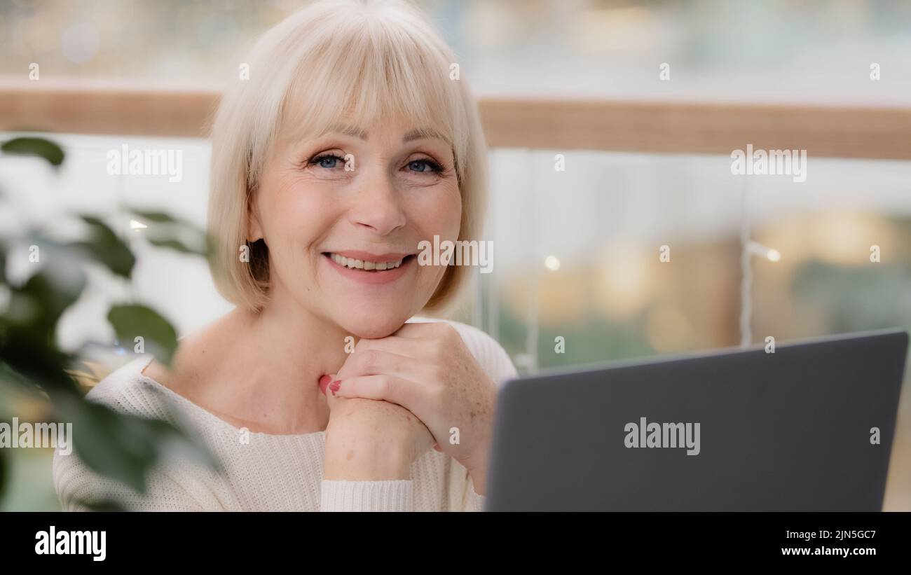 Happy calm mature middle aged woman sitting resting relaxing holding hands under chin looking at laptop screen browsing photos remembering past dreams Stock Photo