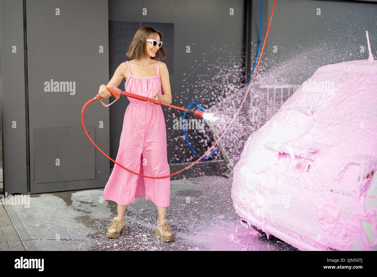 Young woman in pink dress washing her tiny car with nano foam at car wash during summer time. Easy and beautiful self-service at car wash concept Stock Photo
