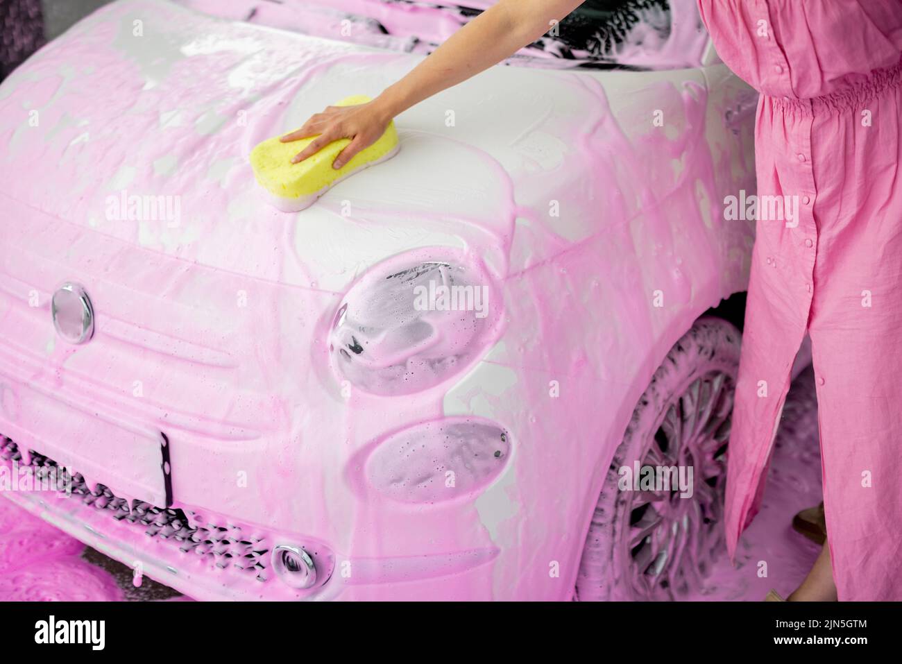 Female person wiping car covered in pink nano foam with yellow sponge at car wash. Close-up view on car hood Stock Photo