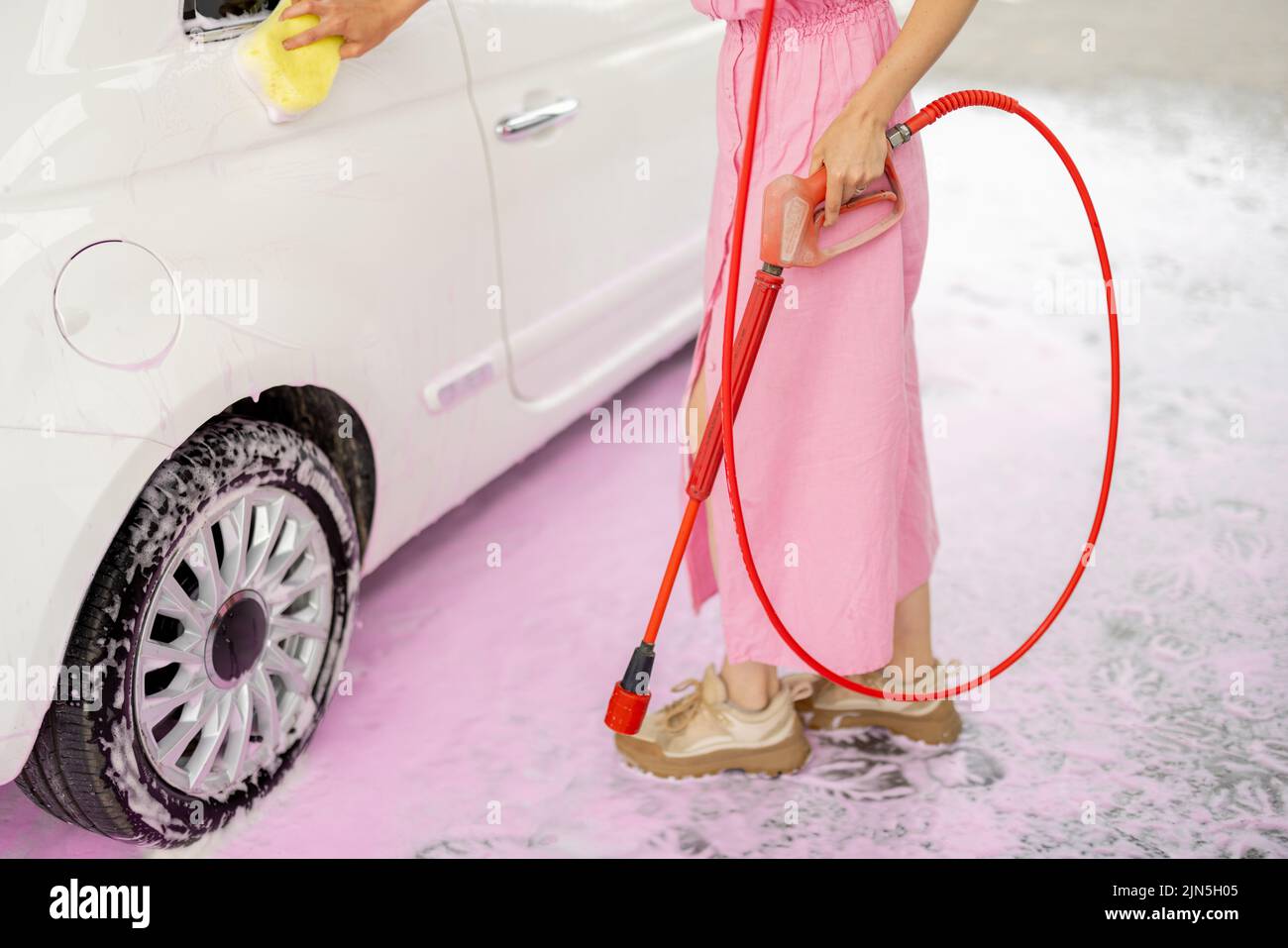 Person wiping car with sponge while washing car with nano foam during summer time. Woman dressed in pink hlding washing gun Stock Photo