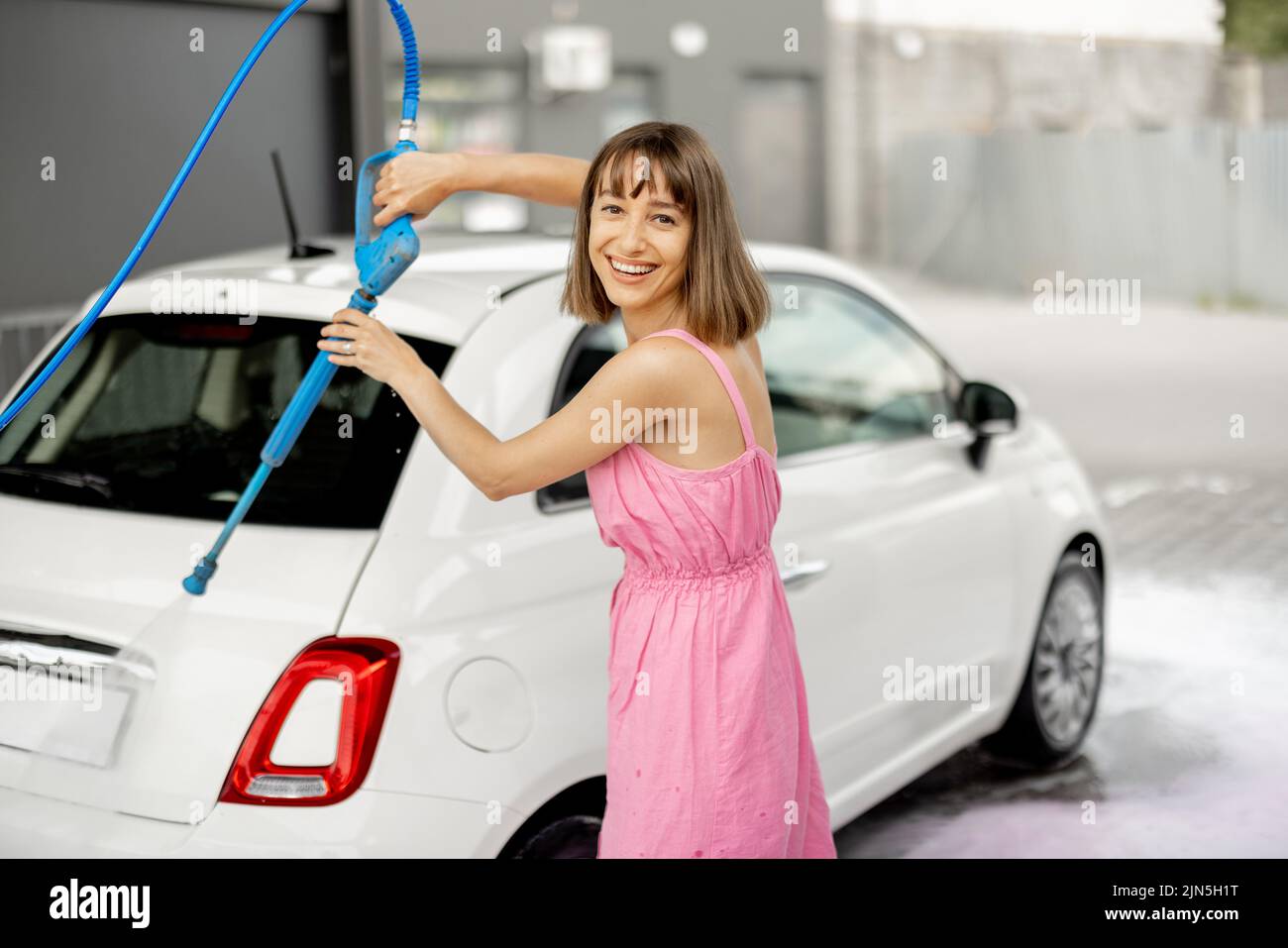 Young woman in pink dress washing her tiny vehicle with washing gun under the pressure at self-service car wash. Easy process of car care by female co Stock Photo
