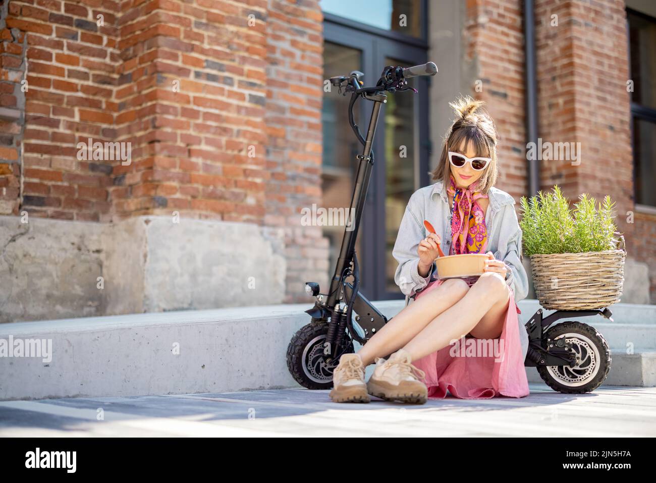 Young stylish woman has a coffee break, eating some take away food while sitting on electric scooter with green plant outdoors. Modern and sustainable Stock Photo