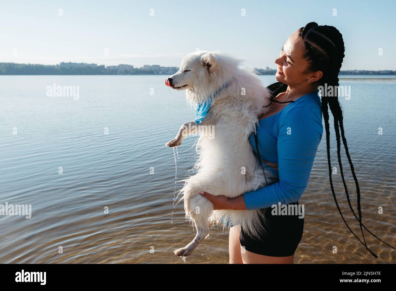 Happy Woman with Dreadlocks Holding on Hands Her Snow-White Dog Japanese Spitz on the Lake at Sunrise Stock Photo