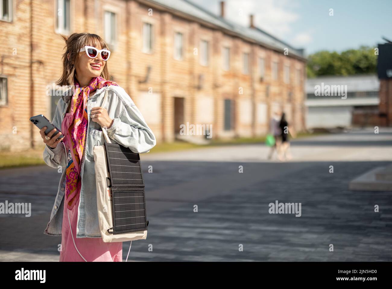 Young stylish woman walks with smart phone and charge it from portable solar panel hanging on bag. Concept of modern and sustainable lifestyle Stock Photo