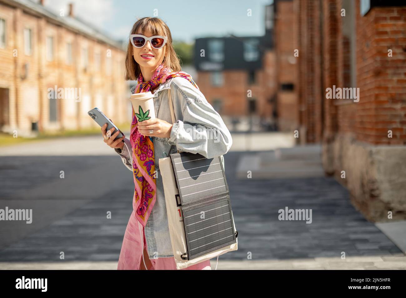 Young stylish woman walks with coffee and smart phone charging it from portable solar panel hanging on bag. Concept of modern and sustainable lifestyl Stock Photo
