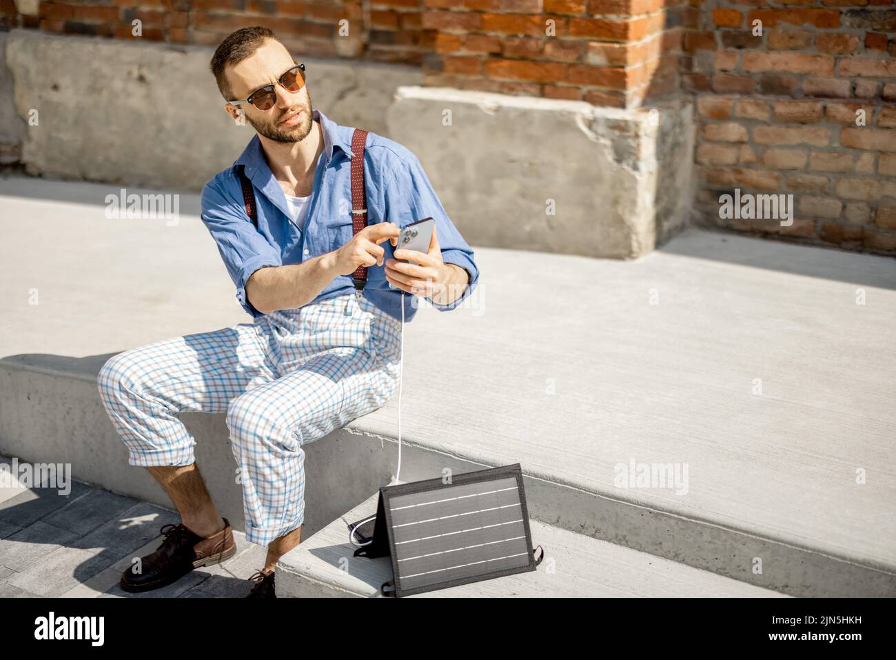 Stylish man use smart phone and charge it from portable solar panel while sitting on street outdoors. Modern sustainable lifestyle and renewable energ Stock Photo