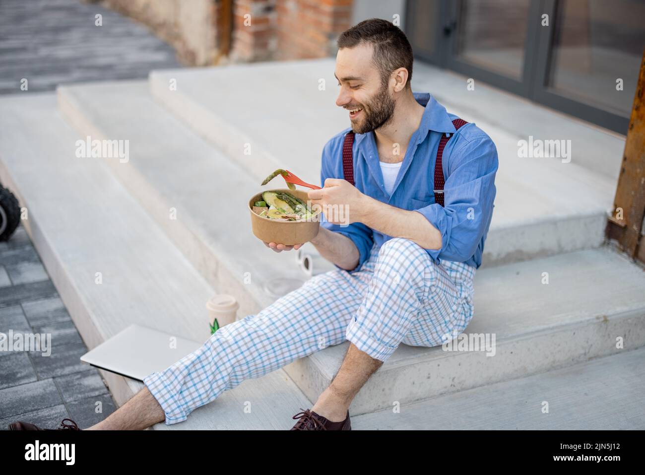 Stylish guy eats healthy take away food from a bowl, while sitting on a street during a lunch time. Concept of healthy vegan lifestyle on work Stock Photo