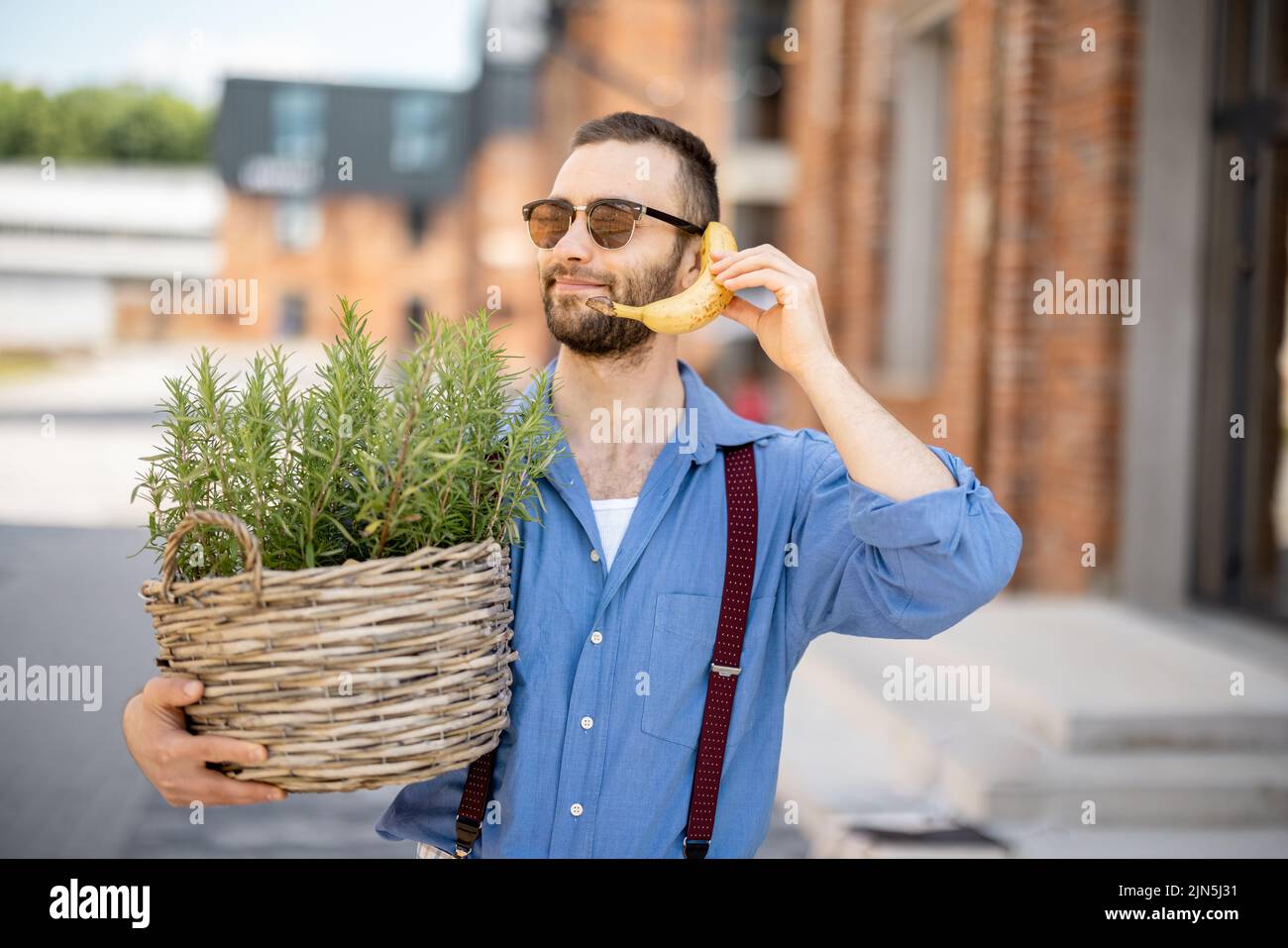 Portrait of weird stylish businessman holds green plant and banana near his ear, like talking on phone outdoors. Cool guy wearing blue shirt and suspe Stock Photo