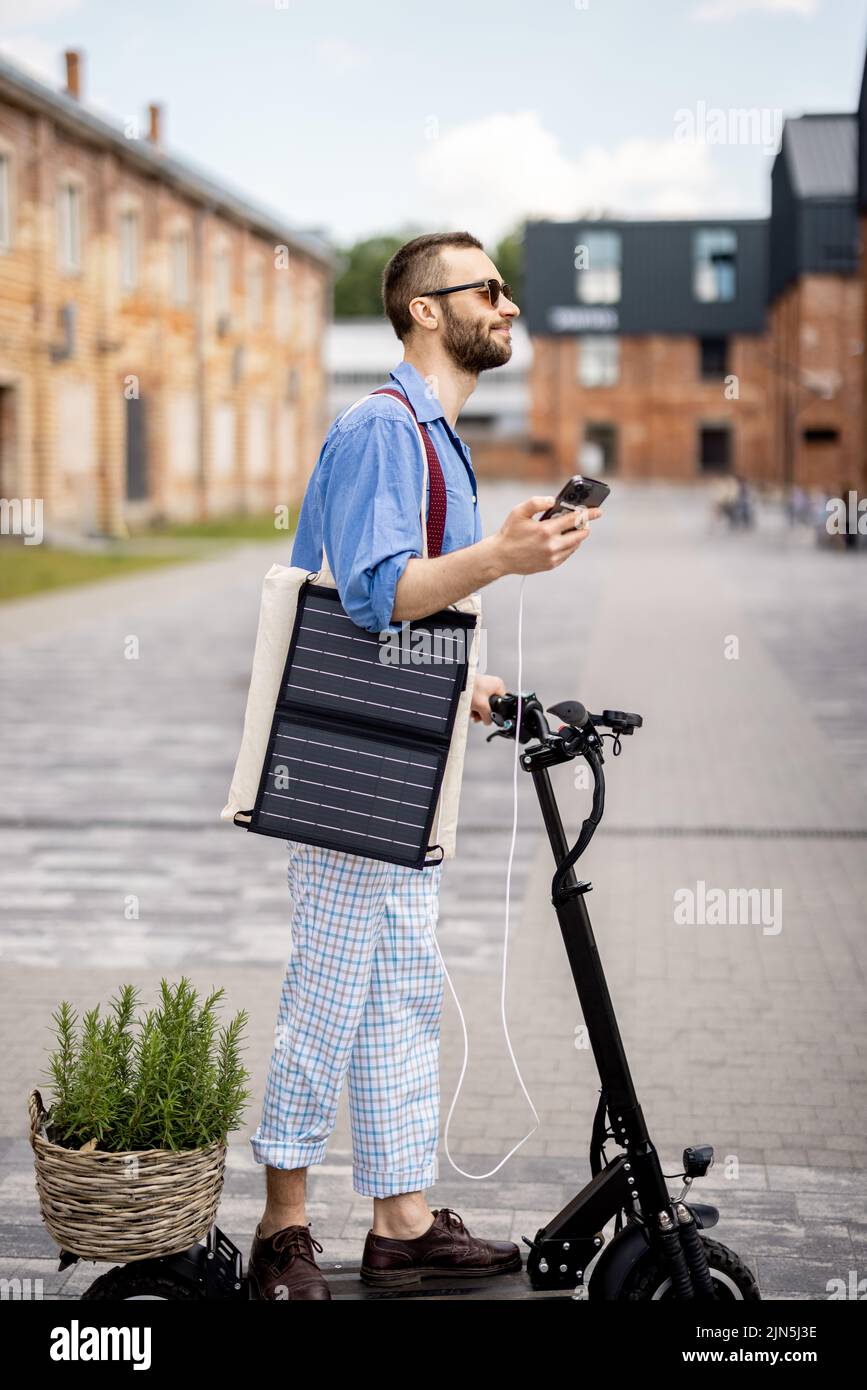 Stylish guy drives electric scooter and charges phone with solar panel on the move on a street at office district. Sustainable business lifestyle and Stock Photo