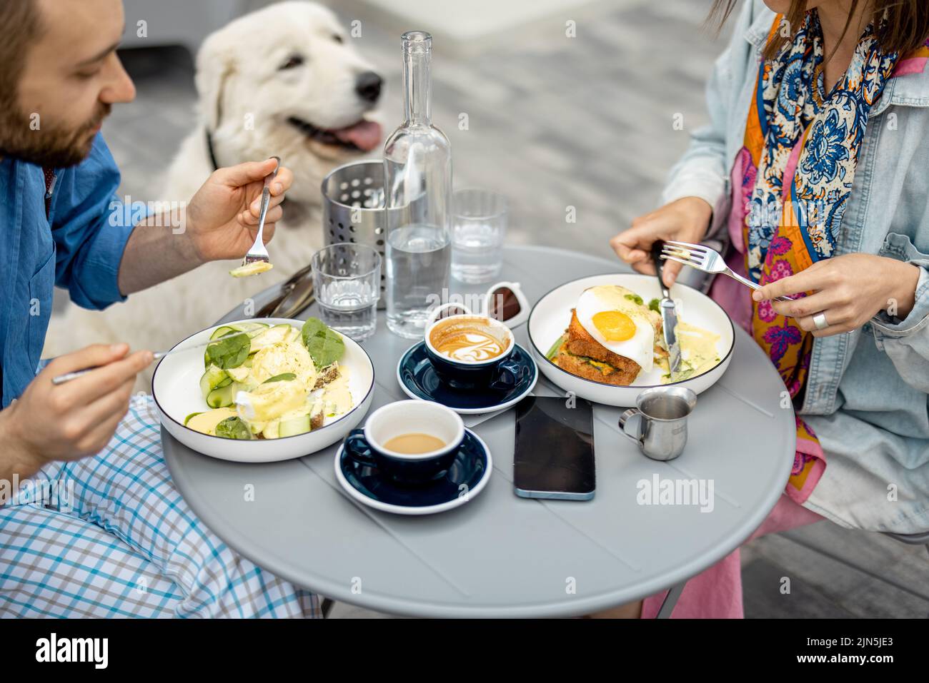 Stylish man and woman have a breakfast while sitting with dog at cafe outdoors Stock Photo