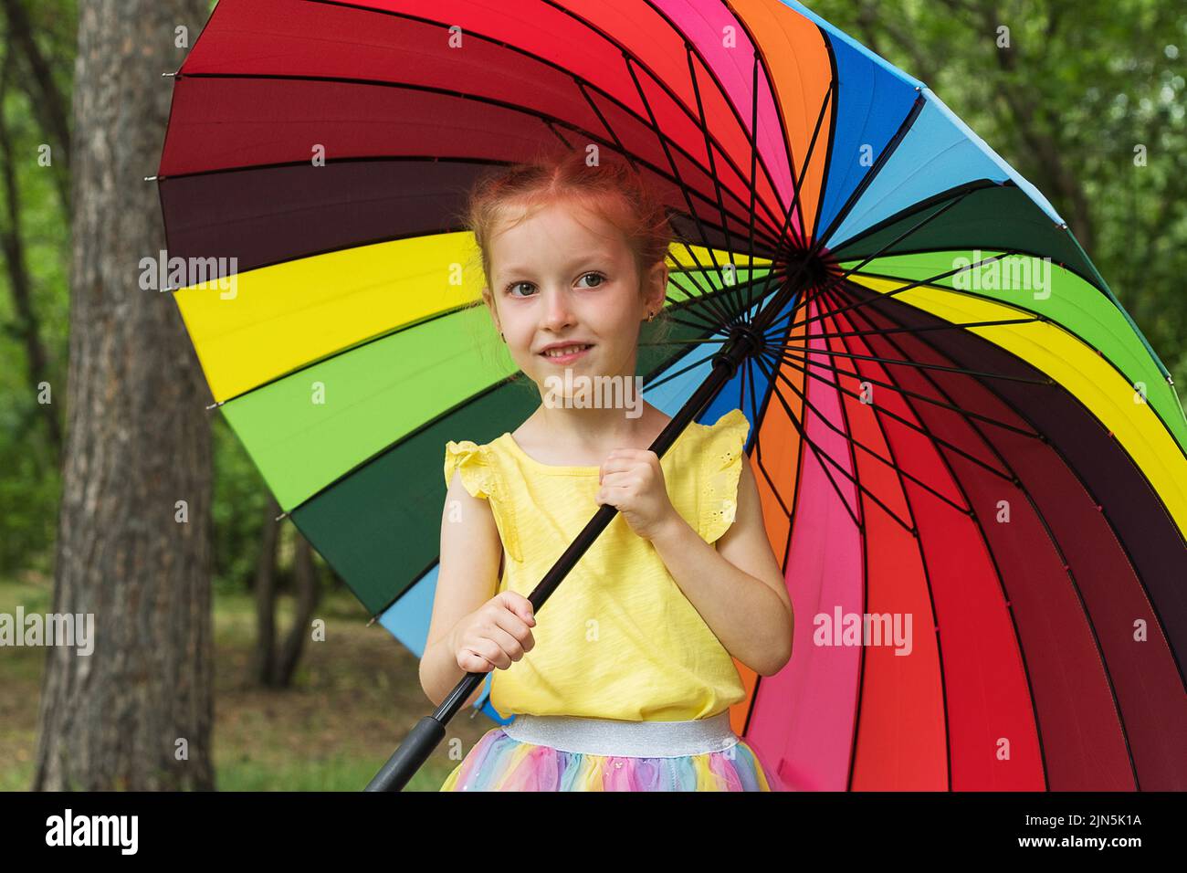 Happy funny girl holding rainbow umbrella. Cute Happy schoolgirl playing in rainy summer park. Kid walking in autumn shower. Outdoor fun by any Stock Photo