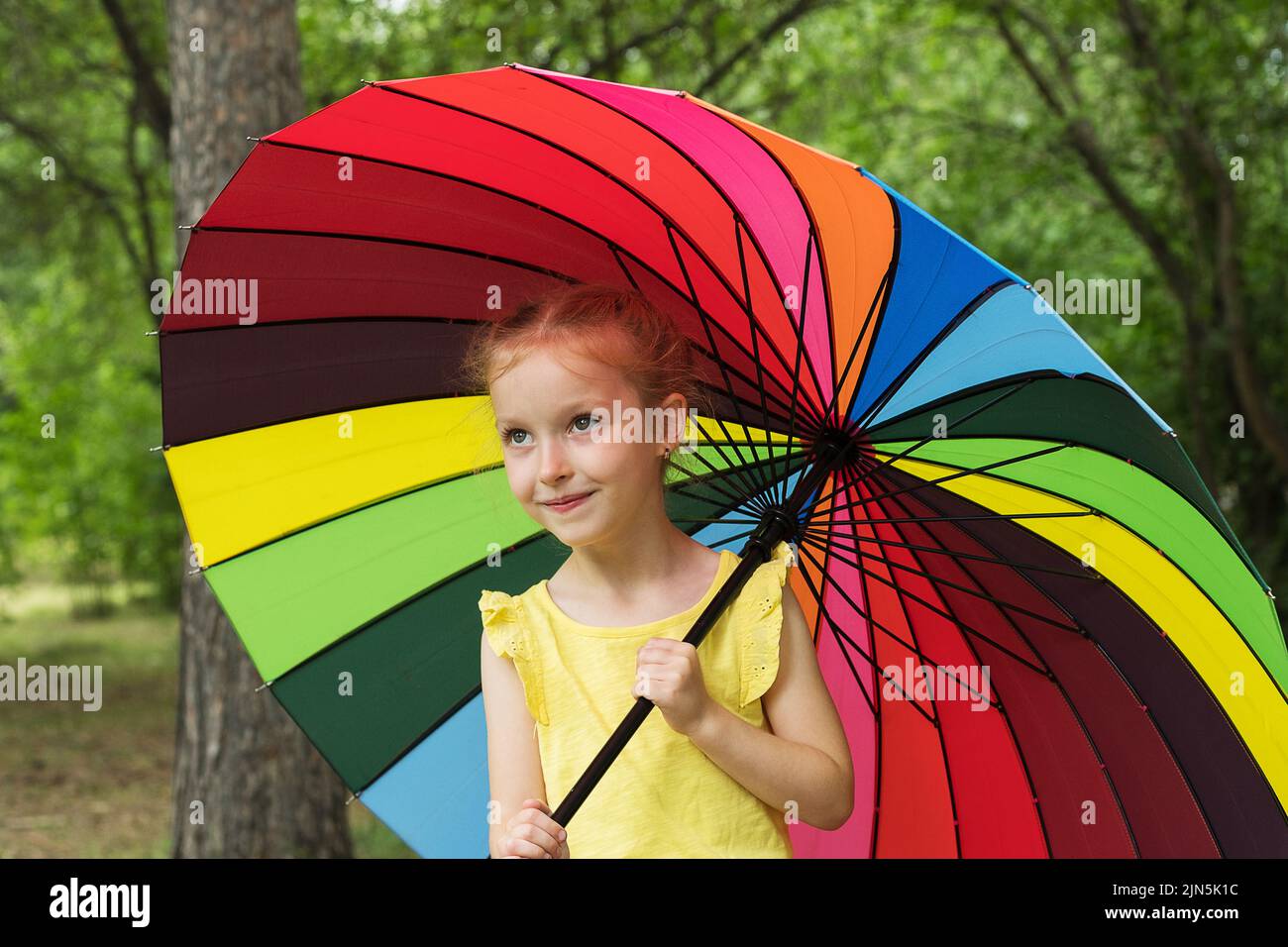 Happy funny girl holding rainbow umbrella. Cute Happy schoolgirl playing in rainy summer park. Kid walking in autumn shower. Outdoor fun by any Stock Photo