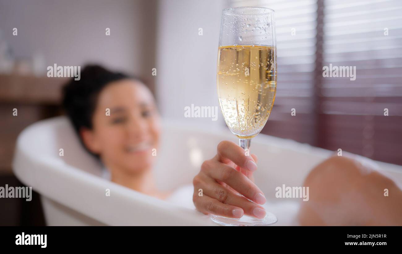 Out of focus young happy relaxed woman lying in foam bath holding glass champagne funny playful lady blowing on foam in bathroom enjoying flavorous Stock Photo