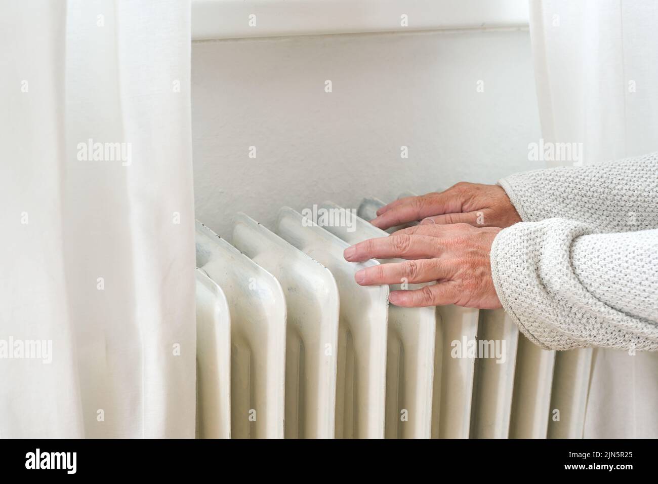 Hands of an elderly woman in woolen clothes feeling low temperature on an old heater, suffering from inflation and rising energy prices, copy space, s Stock Photo