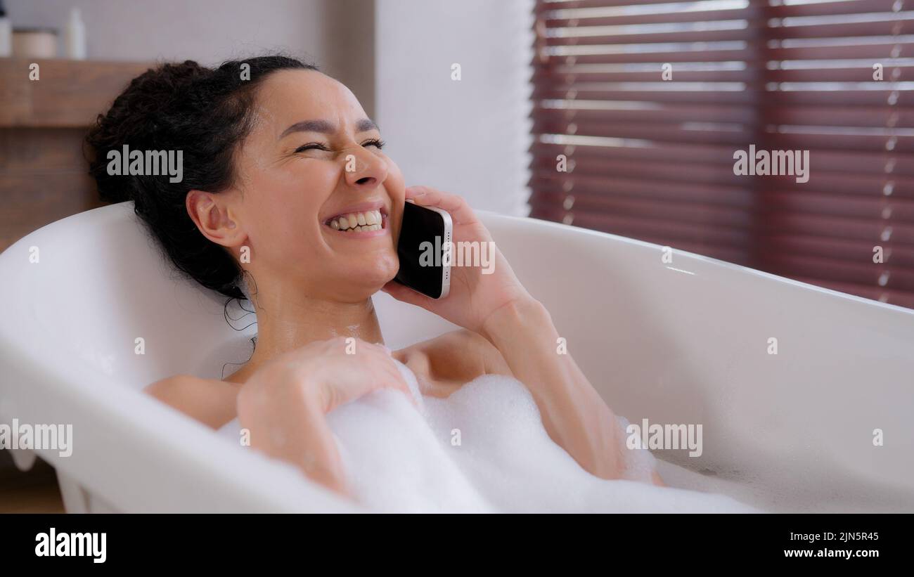 Joyful young woman lies in foam bath relaxes in luxurious bathroom satisfied attractive girl speaks on phone answers friendly call laughs chatting Stock Photo