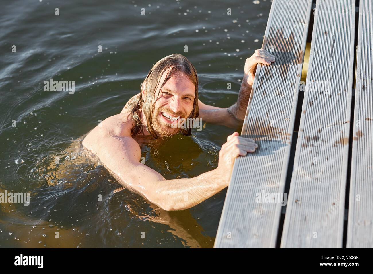Portrait of long haired young man smiling at camera while swimming in lake lit by sunlight Stock Photo