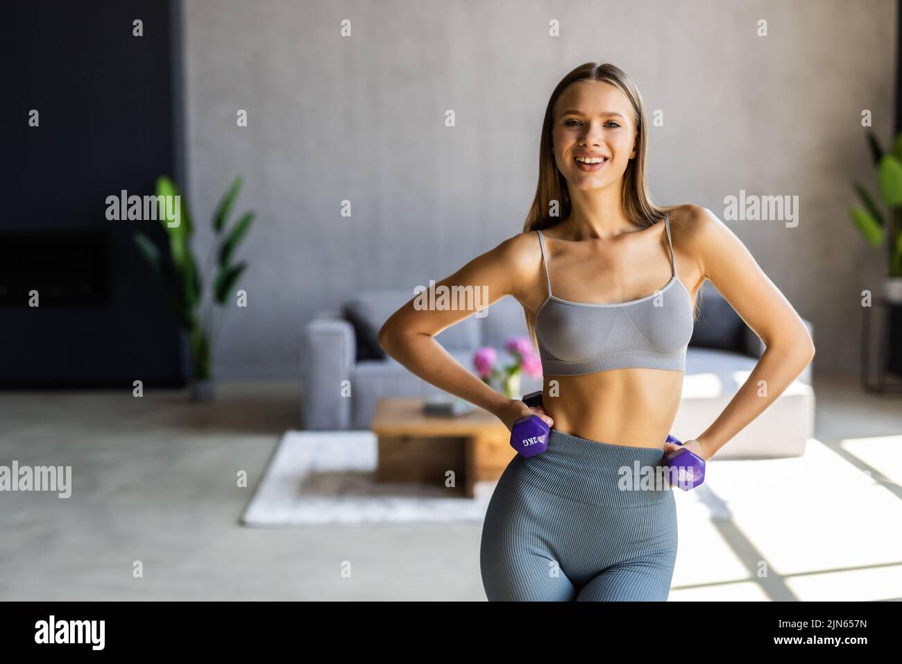 Beautiful and slender young woman performs sports exercises at home using dumbbells. Smiling girl in comfortable sportswear sitting down slightly and Stock Photo