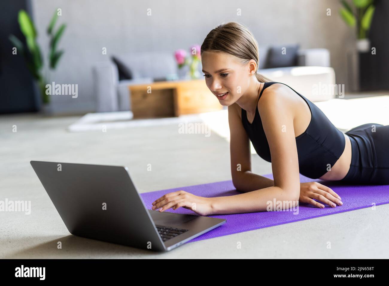 Training Concept. Smiling fit young woman in sports clothes watching fitness videos on internet using laptop near window, lying on floor yoga mat in t Stock Photo