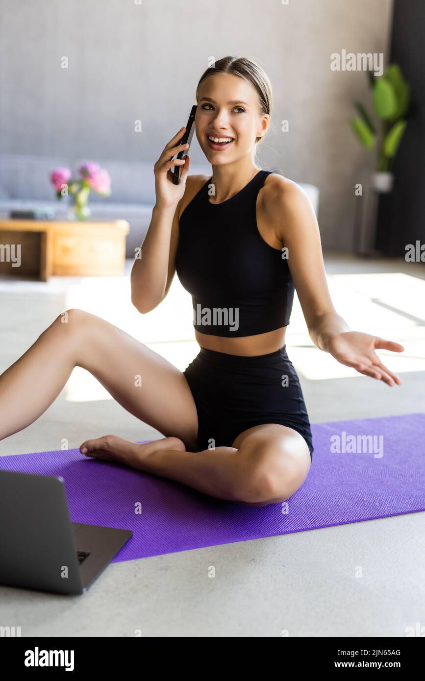 Woman in fitness clothing sitting at home on floor mat, resting after workout, checking mobile phone sport application Stock Photo