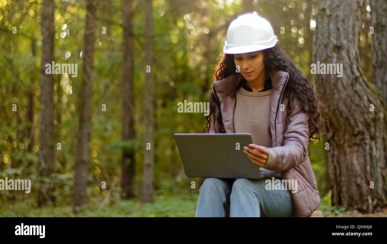 Attractive woman forestry engineer in protective helmet enters data into laptop takes reforestation action young experienced female specialist Stock Photo