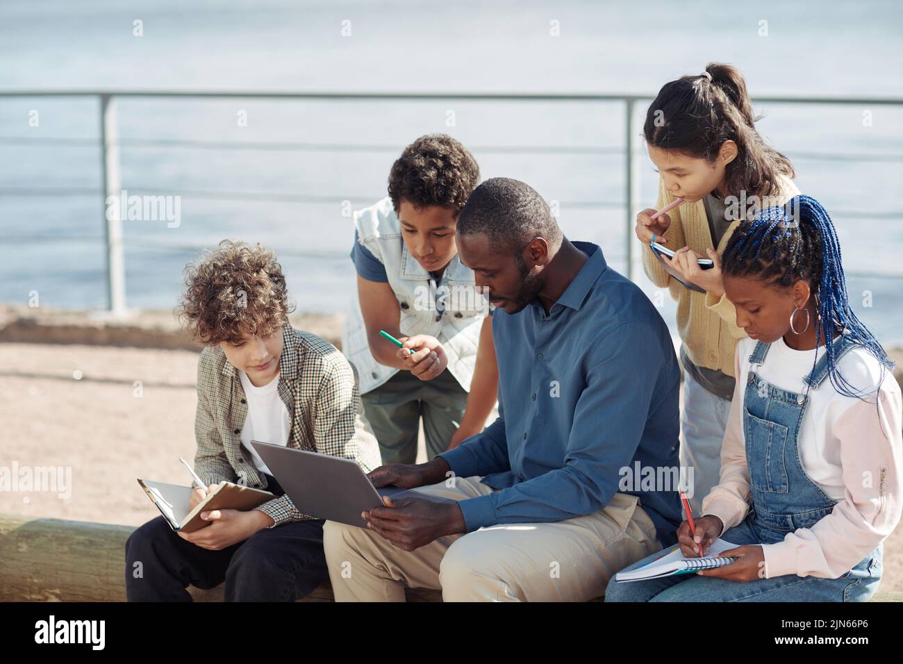 Portrait of diverse group of kids with male teacher using laptop outdoors during Summer school lesson, copy space Stock Photo