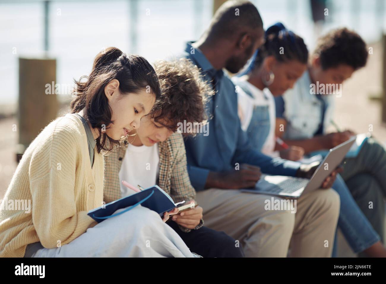 Diverse group of kids sitting in row and writing in notebooks outdoors during Summer school lesson Stock Photo
