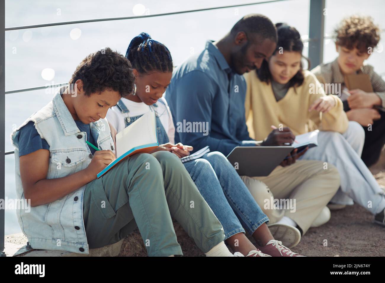 Diverse group of children sitting in row and writing in notebooks outdoors during Summer school lesson Stock Photo