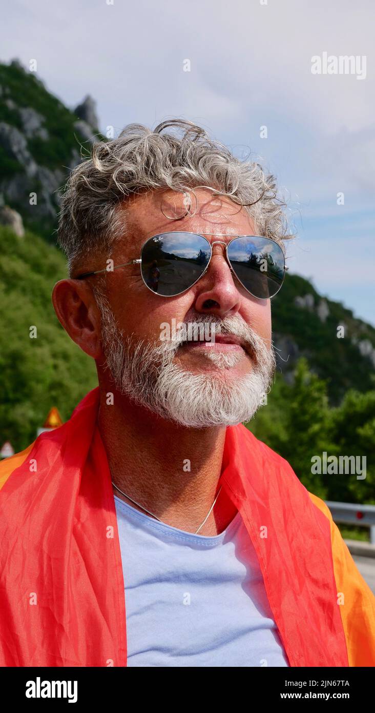Portrait of a gray-haired senior elderly Caucasian man bisexuality with a beard and sunglasses with a rainbow LGBT flag in mountains. Celebrates Pride Month, Rainbow Flag Day Stock Photo