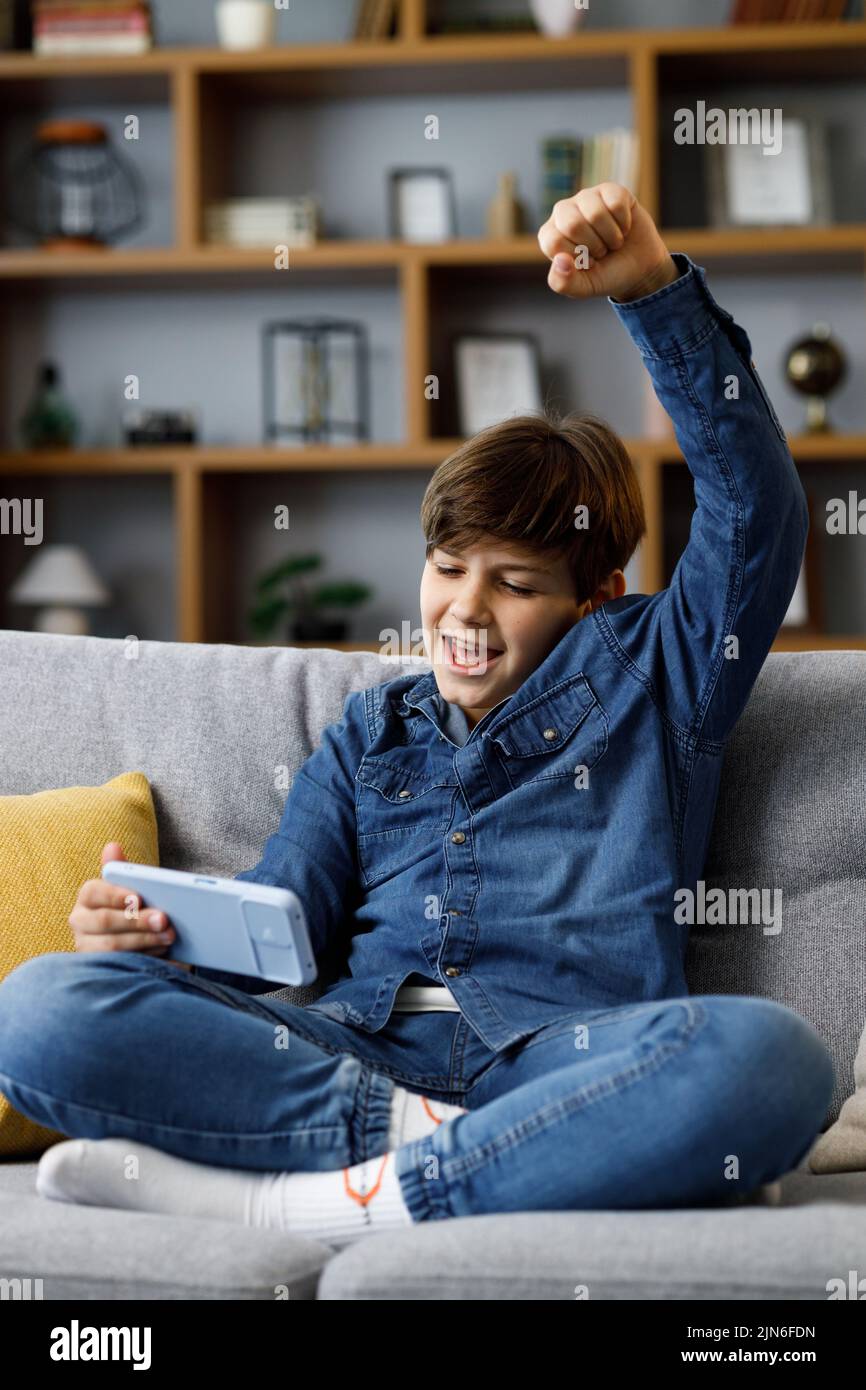 Teens and gaming addiction. Young boy playing video game on smartphone sitting on sofa at home. Cute teenager celebrating victory in online game on Stock Photo