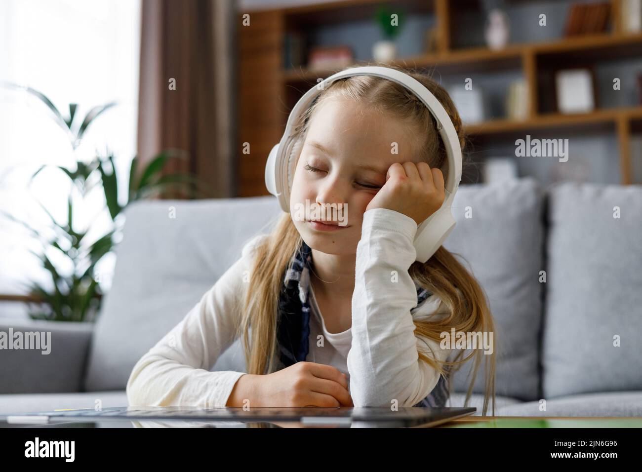 Cute child studying remotely at home using laptop. Tired primary schoolgirl falling asleep during an online lesson with teacher. Distance learning is Stock Photo