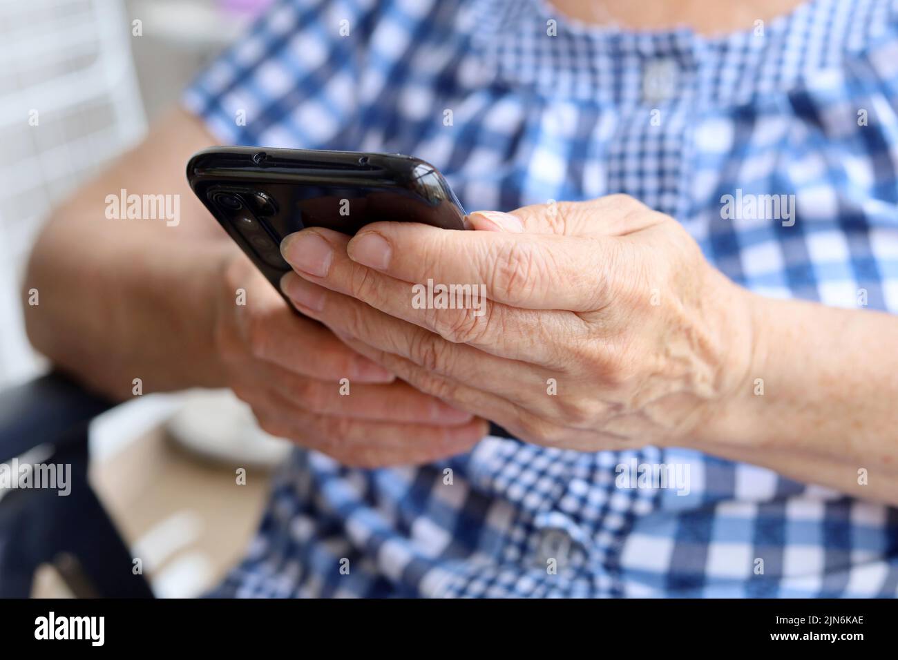 Mobile phone in wrinkled female hands closeup, elderly woman in checkered dress sitting with smartphone. Concept of online communication in retirement Stock Photo