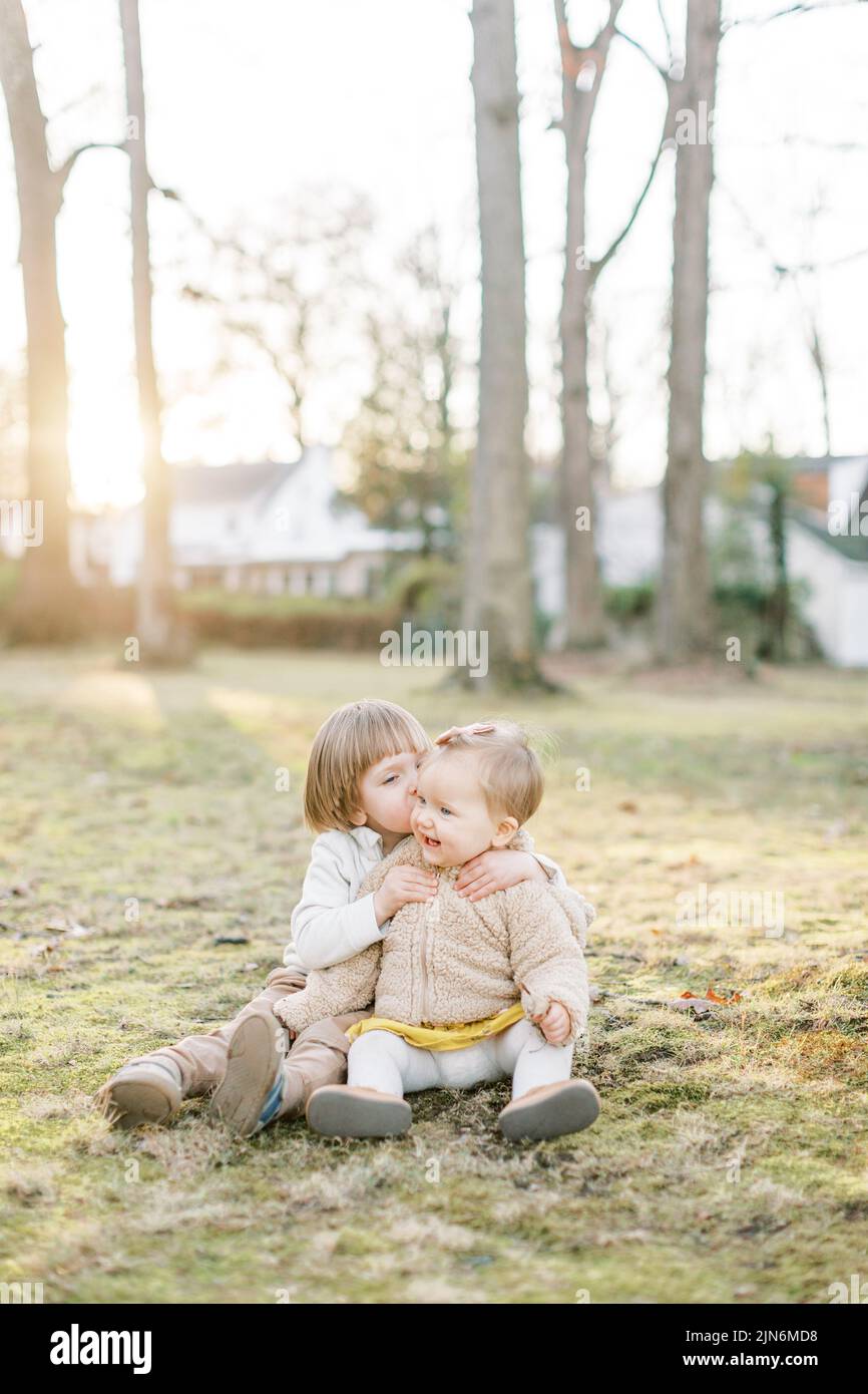 Toddler boy kisses baby sister on cheek while sitting in a field Stock Photo