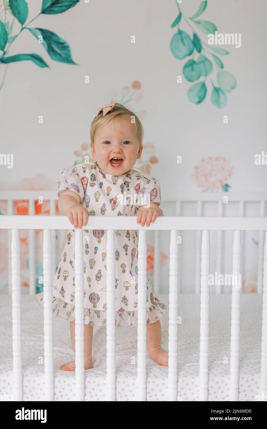 One year old girl in pink dress stands in crib and smiles Stock Photo