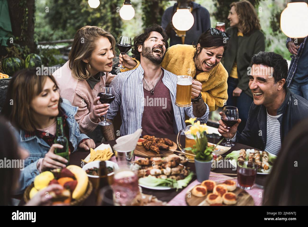 Multiracial group of hilarious friends have fun drinking beer and wine and eating together on the weekend at the restaurant table in the countryside Stock Photo