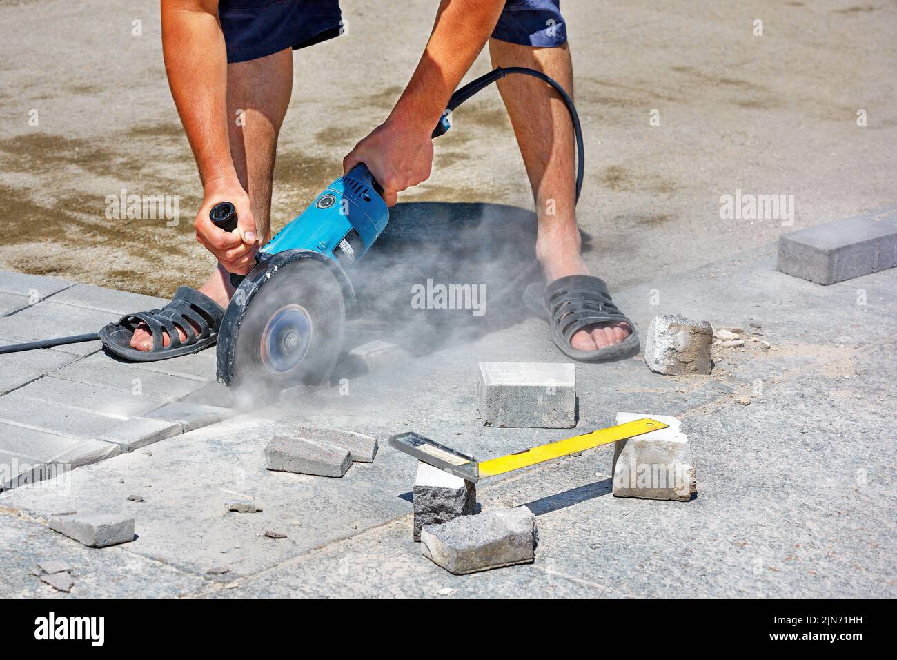 A worker using an electric grinder and a diamond cutting blade cuts paving slabs while laying pavement on a hot afternoon. Copy space. Close-up. Stock Photo