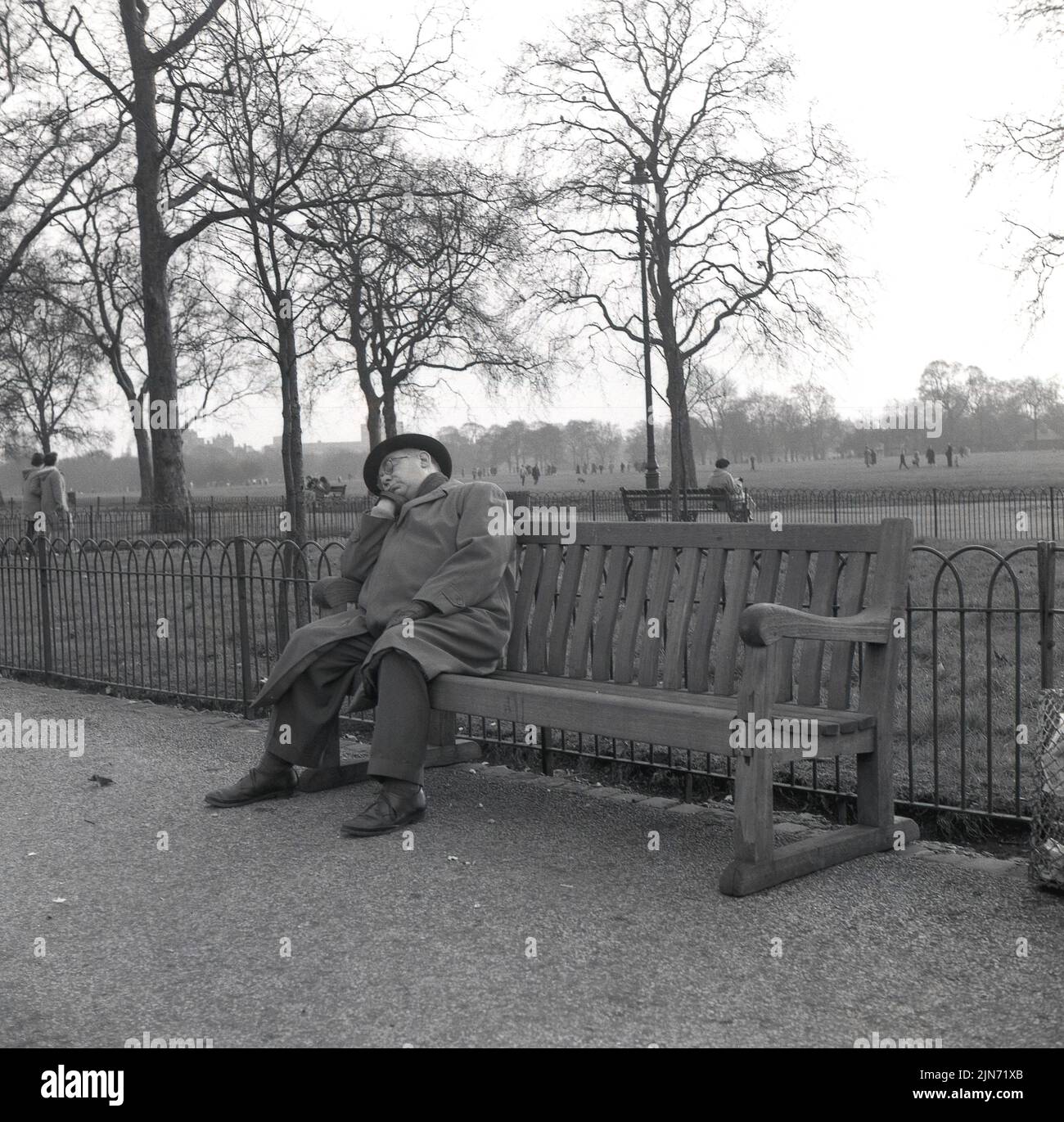 1950s, historical, a gentleman wearing a coat and hat, having a knap on a wooden bench in London's Hyde Park, a large Royal park in the centre of the city. Stock Photo
