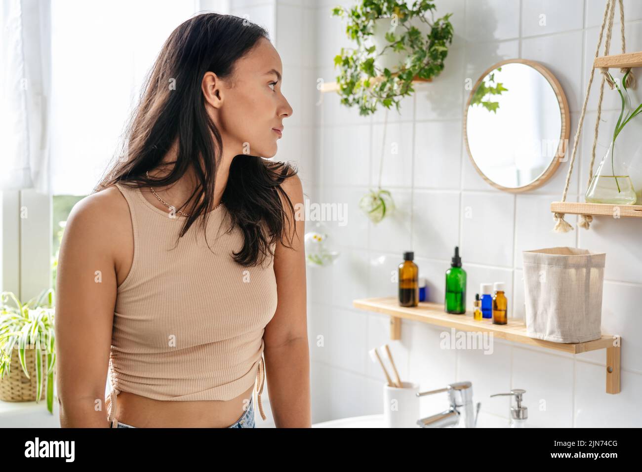 Young beautiful hispanic woman looking into mirror in bathroom. Natural cosmetics, wellness, self-care Stock Photo