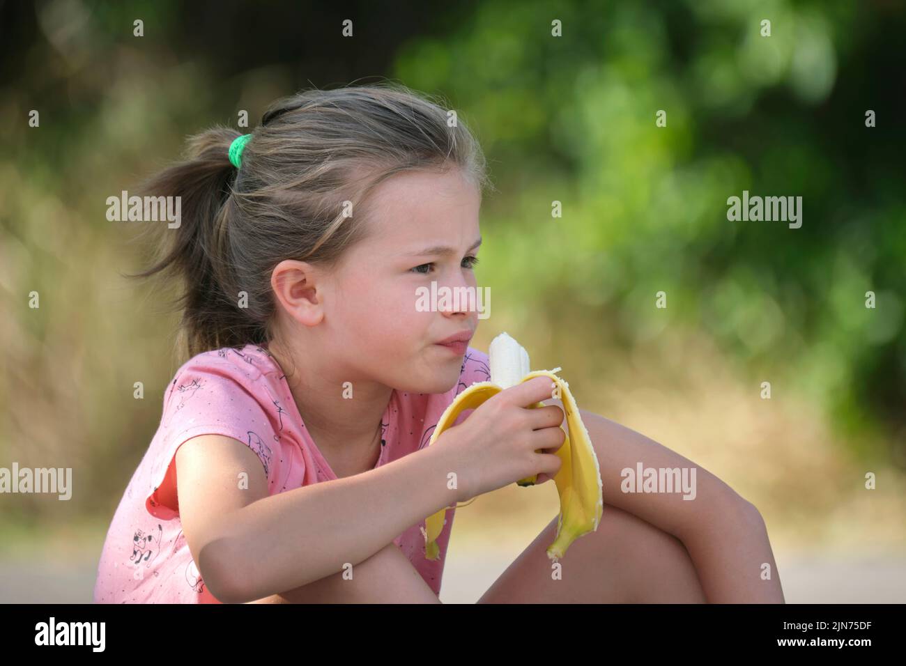 Young pretty child girl eating tasty ripe banana snacking outdoors on summer day Stock Photo