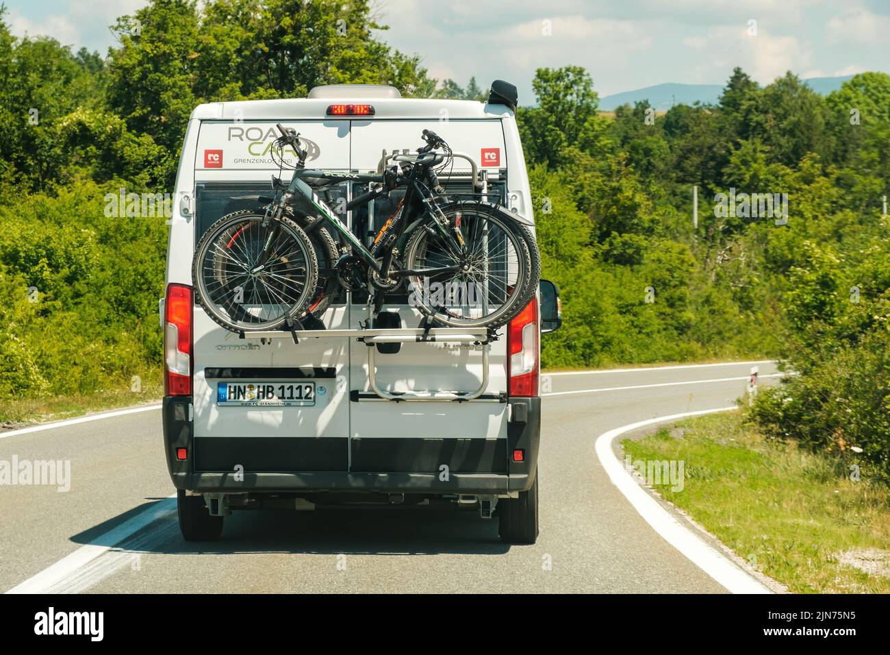 White passenger local bus drives on empty roadway past green trees on sunny day. White vehicle carries fastened sports bicycles on back closeup Stock Photo