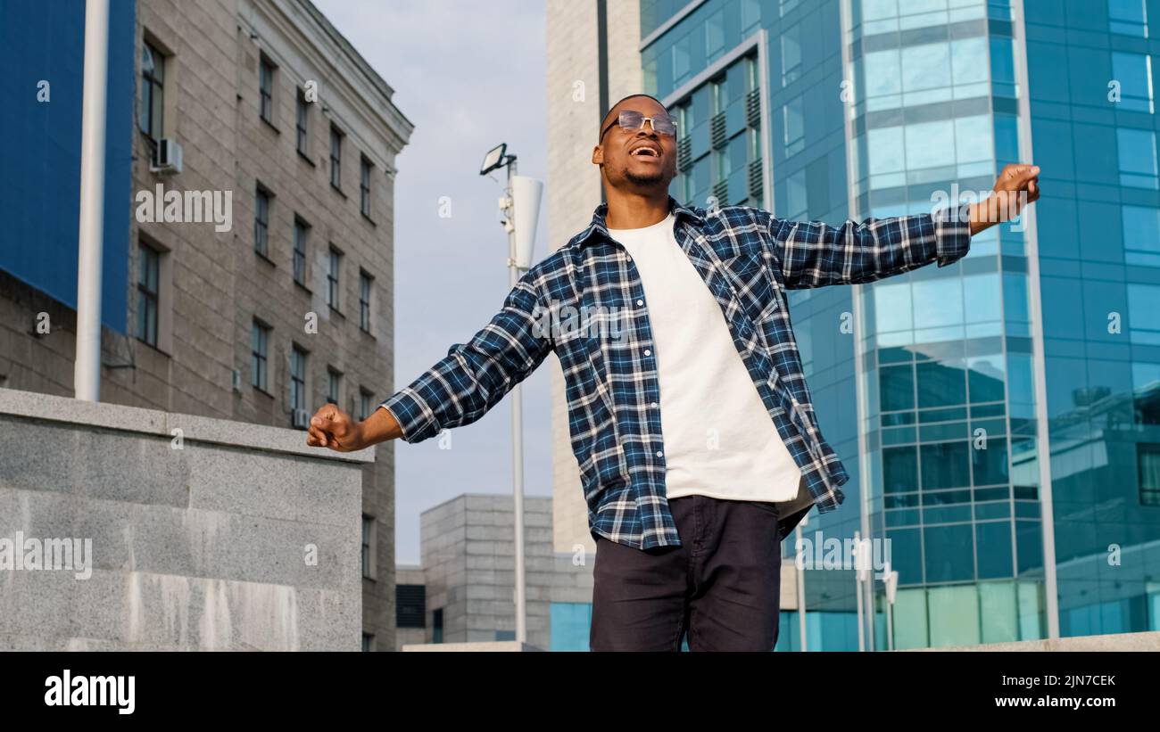 Stylish happy cheerful afro american guy african man singing song sings enjoying music dancing outdoors city building urban background dance having Stock Photo