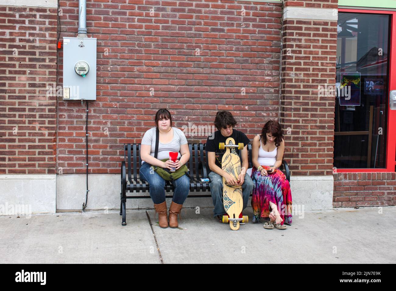 Three young adults sitting on an urban bench - overweight pretty woman - skateboarder with strange expression - girl in tiedyed shirt looking at phone Stock Photo