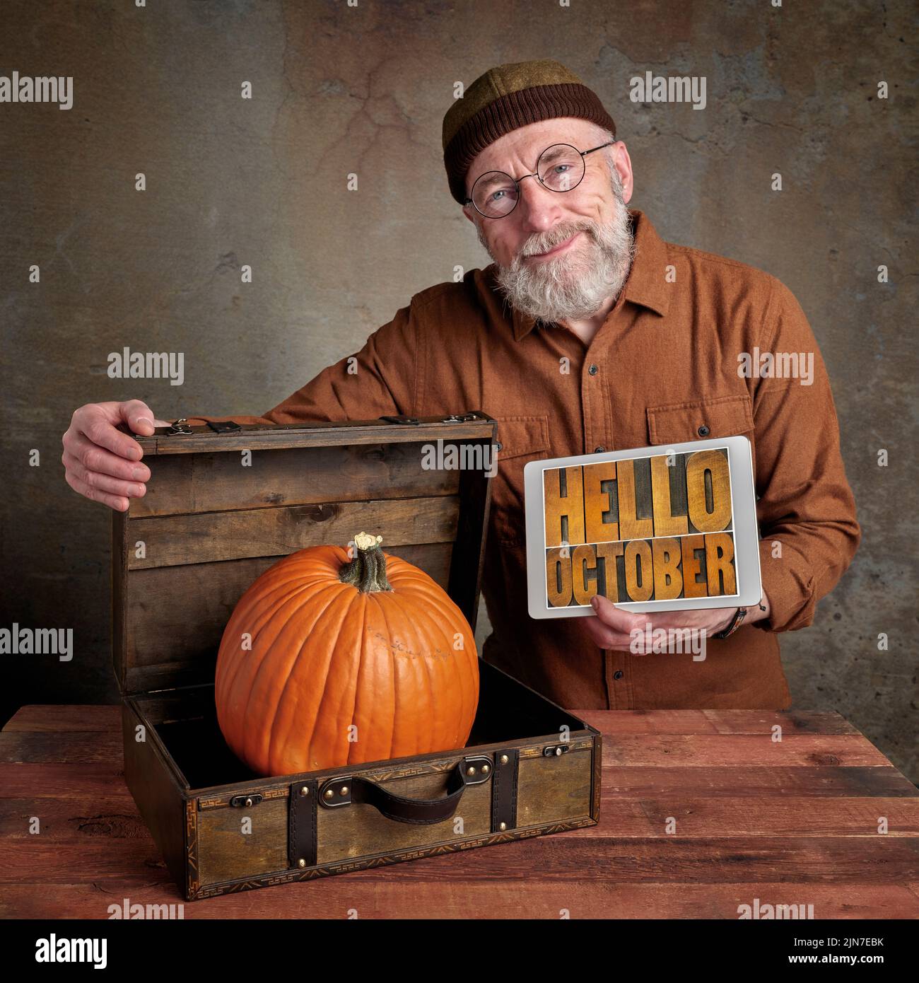 smiling and cheerful senior, bearded man with a big pumpkin in a small retro suitcase and a digital tablet with hello October text in letterpress wood Stock Photo