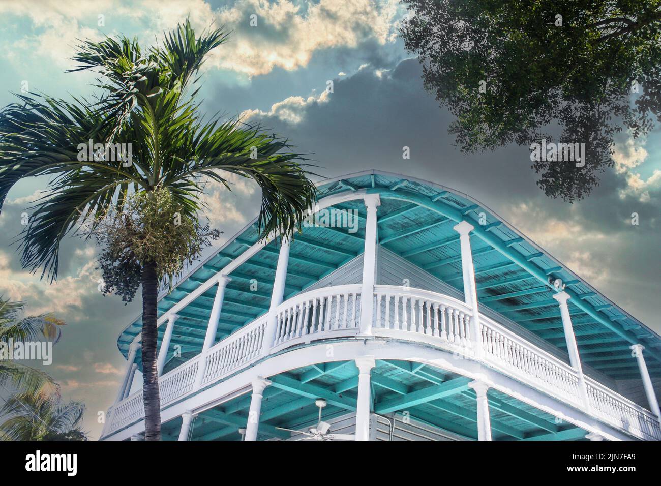 Tropical turquoise building with balcony around it against stormy sky with palm tree - looking up at an angle - perspective Stock Photo