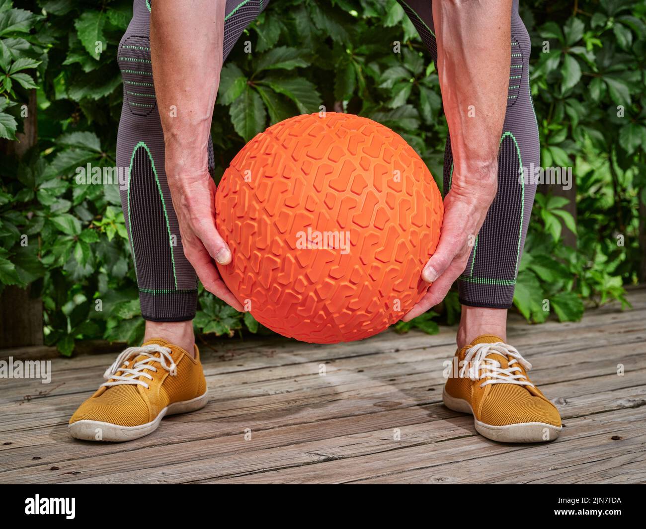 male wearing compression sleeves is exercising with a heavy slam ball on a backyard deck, functional fitness and backyard gym concept Stock Photo