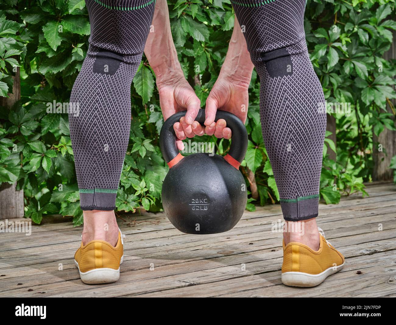 male wearing compression knee and calf braces is exercising with a heavy iron kettlebell on a backyard wooden deck Stock Photo